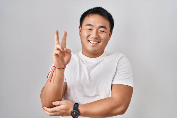 istock Young chinese man standing over white background smiling with happy face winking at the camera doing victory sign with fingers. number two. 1446458432