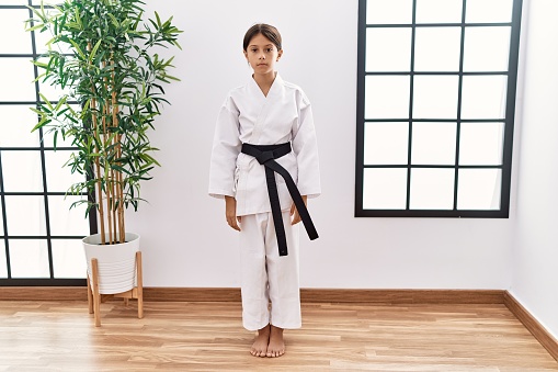 Young hispanic girl wearing karate kimono and black belt relaxed with serious expression on face. simple and natural looking at the camera.