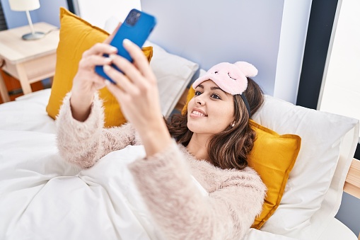 Young beautiful hispanic woman wearing sleep mask make selfie by smartphone at bedroom