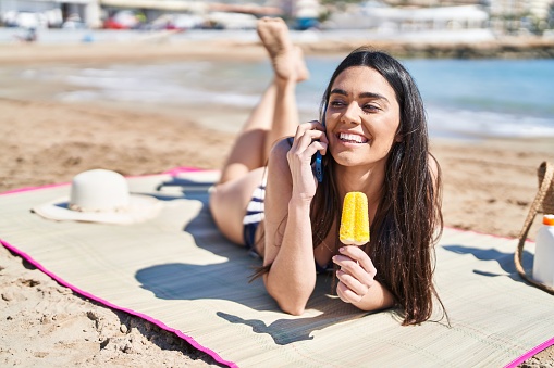 Young hispanic woman talking on the smartphone eating ice cream lying on sand at seaside