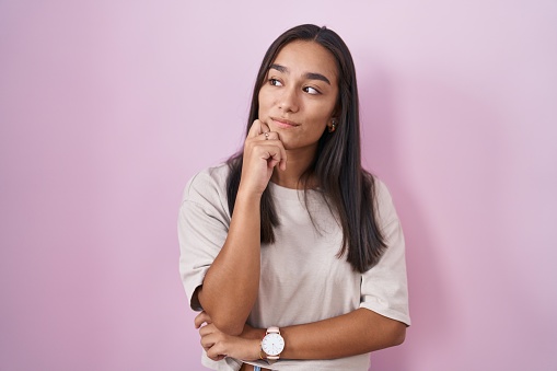 Young hispanic woman standing over pink background with hand on chin thinking about question, pensive expression. smiling with thoughtful face. doubt concept.