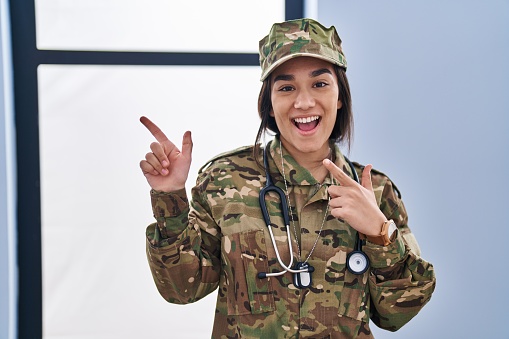 Young south asian woman wearing camouflage army uniform and stethoscope smiling and looking at the camera pointing with two hands and fingers to the side.