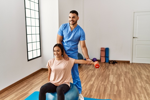 Latin man and woman wearing physiotherapist uniform having rehab session using fit ball and dumbbell at rehab center