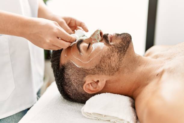 young hispanic man having facial mask treatment at beauty center - masker stockfoto's en -beelden