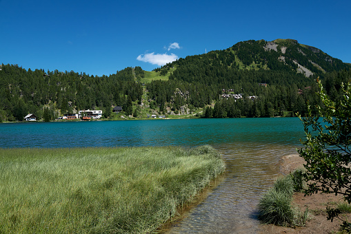 A small stream feeds a greeen shimmering mountain lake, which is surrounded by wooded mountains with a few houses.
