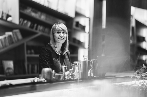 Black and white shot of smiling waitress standing behind the counter and working on laptop in the evening