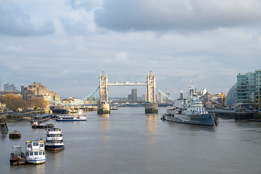Elevated view over water of north and south shores, warship, and iconic 19th-century bascule and suspension bridge connecting Tower Hamlets and Southwark.