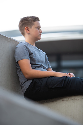 Young thoughtful teenage boy sitting in school backyard on a concrete wall, enjoying break time.
Boy with dark blonde hair wearing gray t-shirt and black pants looking curiously into the distance. 
Background is out of focus, modern concrete contemporary building with trees.