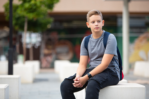 Young serious and worried school teenage boy sitting on a concrete cube, curiously looking in the distance. \nBoy with dark blonde hair and blue eyes wearing gray t-shirt, black pants, backpack and a black smart watch. \nBackground is out of focus, modern concrete with contemporary details and trees.