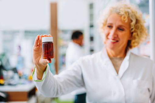 A  Senior Caucasian pharmacist is at the drugstore holding a bottle of medicine and looking at the camera.