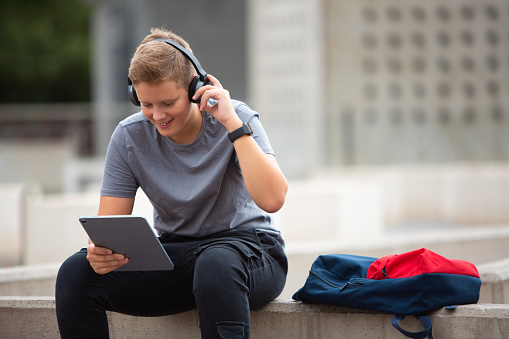 Young smiling teenage boy sitting, leaning on his elbow, wearing headphones and holding tablet. 
Boy with dark blonde hair wearing gray t-shirt, black pants and a black smart watch adjusting his headphones while looking at his tablet. 
Background is out of focus, modern concrete with contemporary details and trees.