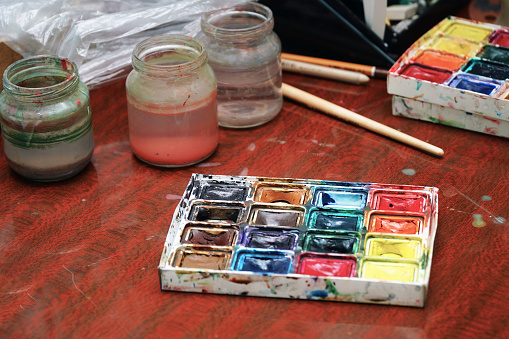 Watercolor paints, a paint brush and jars of water on the table. Selective focus