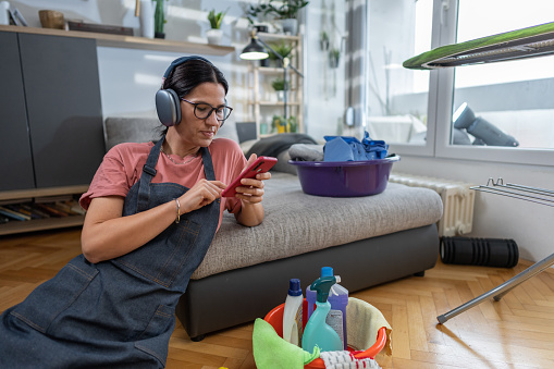 Housewife with wireless headphones using smart phone while sitting on the floor in living room at home.