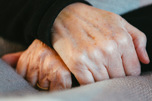 Close up of a senior woman's hands clasped together resting on a blanket. The hands are wrinkled with liver spots.