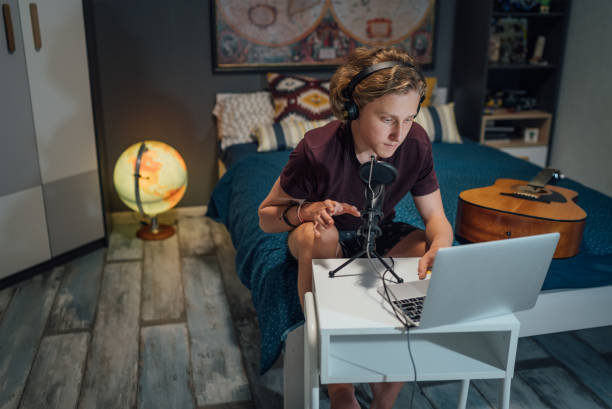 Home sound studio young teenager portrait using laptop computer and Headphones, playing guitar and recording voice music with microphone in the kid's room. Audio recording technology concept image stock photo