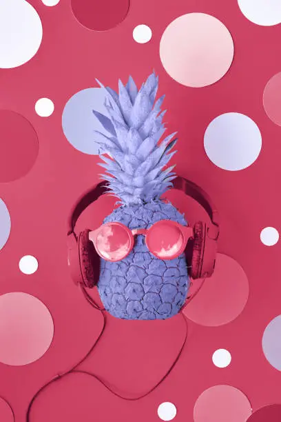 Photo of Viva Magenta color of the year 2023. Funny pineapple painted purple on magenta background with circles. Pineapple character in sunglasses and earphones.