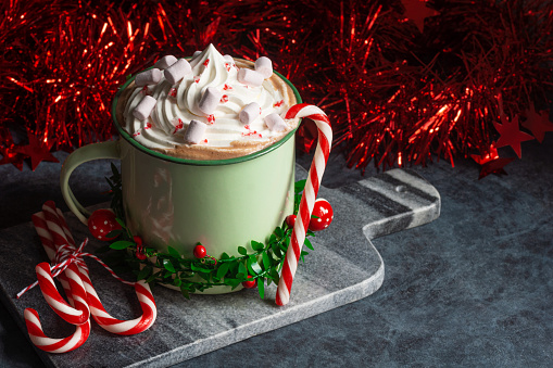 A large green mug filled with peppermint hot chocolate (cocoa) with mint candy canes, a swirl of whipped cream, and pink marshmallows. The hot drink is wrapped in a garland of leaves and berries and has candy canes and tinsel off to the side for Christmas or winter holidays.