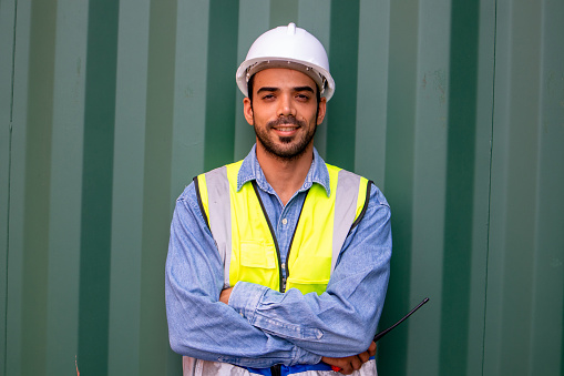 Portrait of confident young hispanic male engineer with container background.