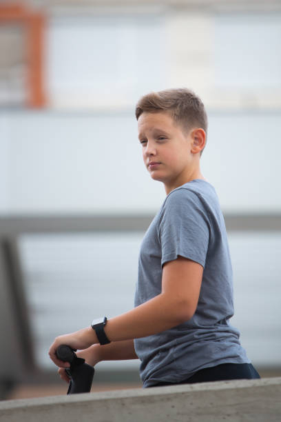 A young teenage boy standing on his scooter behind a concrete wall looking thoughtfully sideways.\nBoy with dark blond hair and blue eyes wearing gray t-shirt, black pants, and a black smart watch. \nBackground is out of focus, modern concrete with contemporary details.