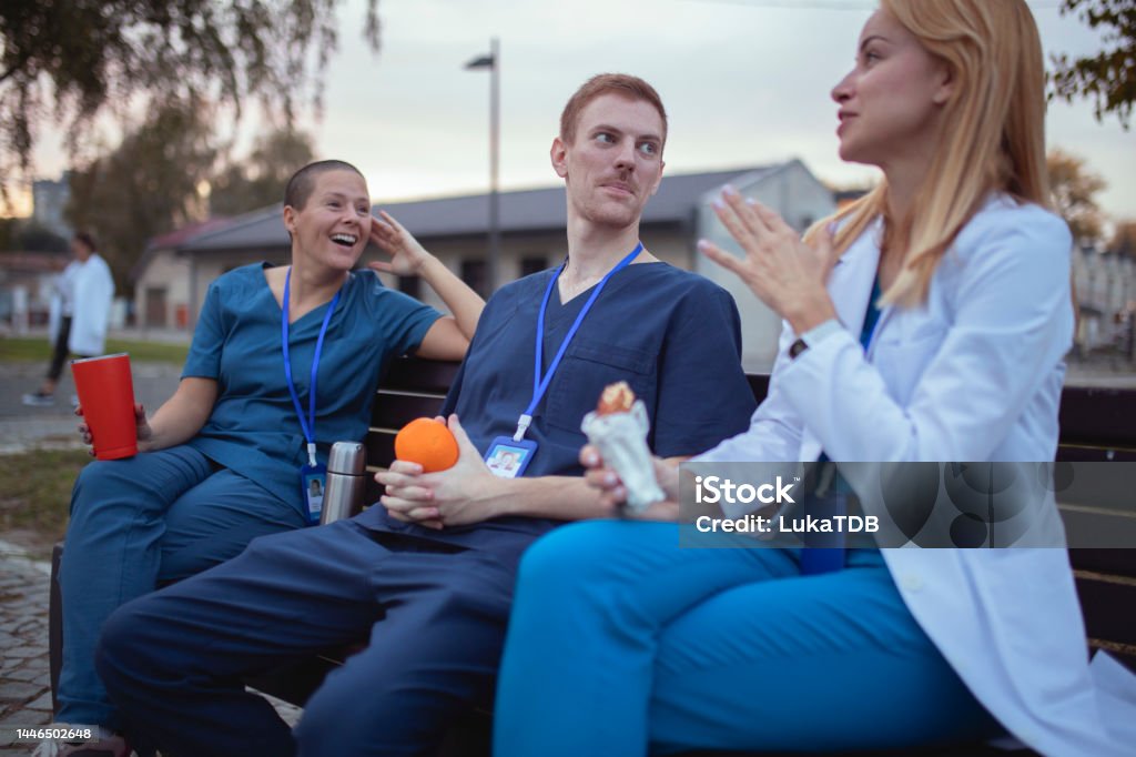 A doctor in a white lab coat talks to her colleagues in uniform, while they sit on a bench A small group of medical workers sits on a bench in the hospital yard and chats during a coffee break Nurse Stock Photo