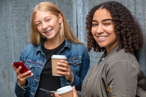 Mixed race friends pretty blonde teenager and biracial African American girl female young woman using their smart phones for social media and drinking takeout coffee outside