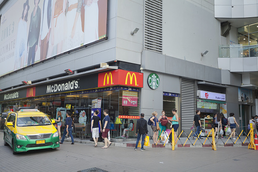 Tourists and people outside of McDonald's amd Starbucks on Phetchaburi Road in Bangkok downtown. Both are between two malls. At left side is a taxi on driveway. There are white and asian tourists and thai people.