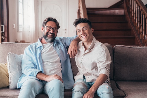 Happy father and son sitting on sofa at home. Cheerful father with teenage son sitting on couch in the living room of apartment. Portrait of father and son spending leisure time at home