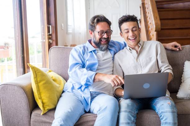 happy father and son looking at laptop screen while sitting on couch in living room at home. father with teenage son browsing social media content using laptop in living room - teenager parent father son imagens e fotografias de stock