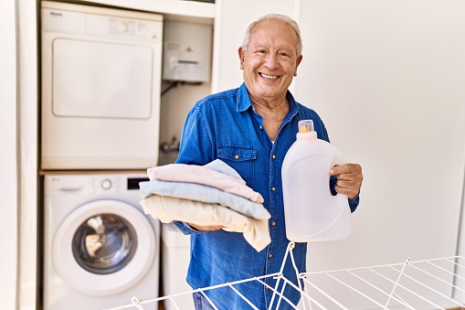 Senior caucasian man holding detergent and folded clothes standing by washing machine at the terrace.
