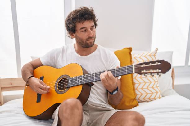 istock Young hispanic man playing classical guitar sitting on bed at bedroom 1446542903