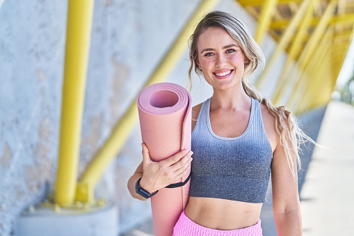Young blonde woman wearing sportswear holding yoga mat at street