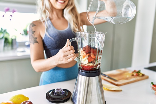 Young woman smiling confident pouring water on blender at kitchen