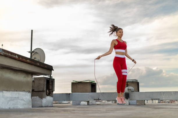 Young woman exercising with skipping rope on rooftop Attractive young woman jumping skipping rope outdoors on building rooftop in city in urban area. She is wearing sports clothing and looks very fit and healthy Skipping stock pictures, royalty-free photos & images