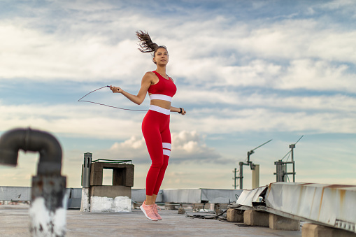 Attractive young woman jumping skipping rope outdoors on building rooftop in city in urban area. She is wearing sports clothing and looks very fit and healthy