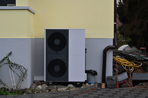 Dark grey colour air source heat pump installed outside the house painted grey and pastel yellow for heating and cooling homes. They work by moving outdoor heat indoors, or vice versa.