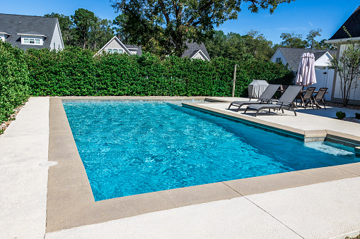A rectangular new swimming pool with tan concrete edges in the fenced backyard of a new construction house