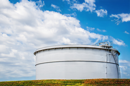 Large white petrochemical storage tank at terminal against blue cloudy sky with colorful grass and red clay foreground