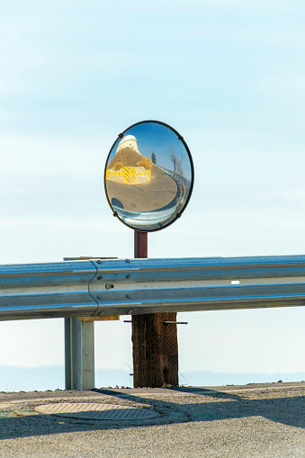 road saftey mirror or street mirror on metal gaurd rail in sun with wooden supports and blue and white sky background late afternoon or early evening