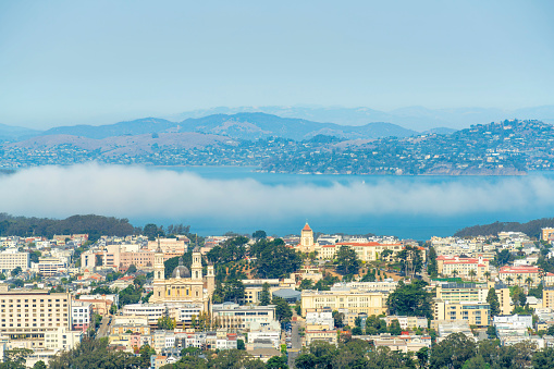 bright multicolored neighborhoods and houses and buildings in san francisco in partial sunlight with moutains and ocean in background with clouds and fog and gray and blue gradient sky late afternoon