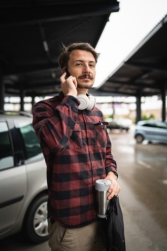 One man young adult caucasian male standing at the parking lot in day using mobile phone smartphone talk while going to work wait alone in day real people copy space hold briefcase and cup of coffee