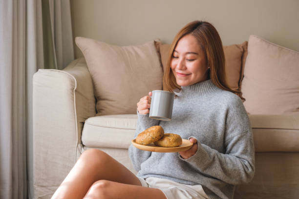 家で熱いコーヒーを飲みながらベーグルの皿を持つ美しいアジアの若い女性のポートレート画像 - model home house home interior plate ストックフォトと画像