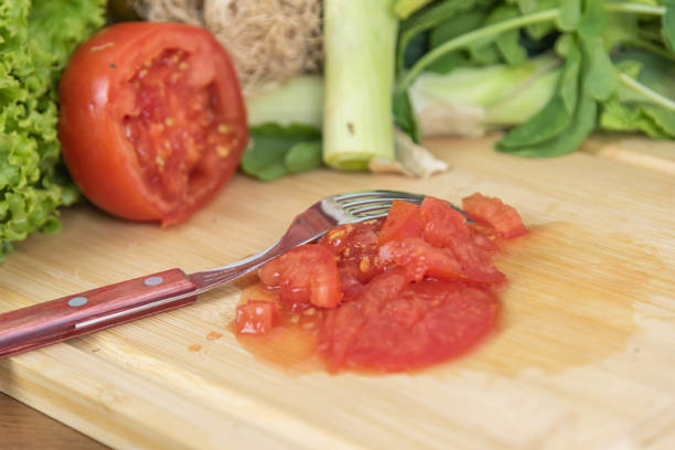 Hand is squeezing and crushing a ripe red tomato, with other squashed tomatoes scattered on green grass, depicting food waste or a playful act