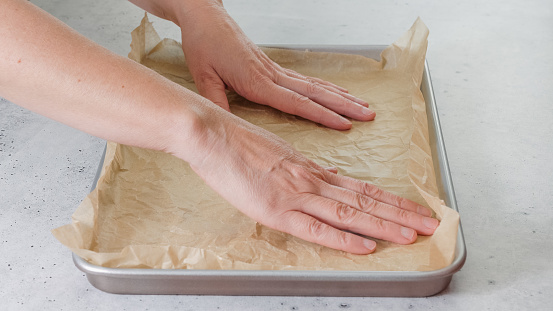 Woman's hands placing parchment paper onto a baking pan, close-up on the kitchen table