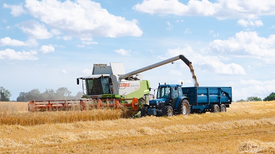 Cambridge, United Kingdom – July 17, 2022: A CLAAS Combine Harvester harvesting a field while emptying its grain into a nearby tractor.