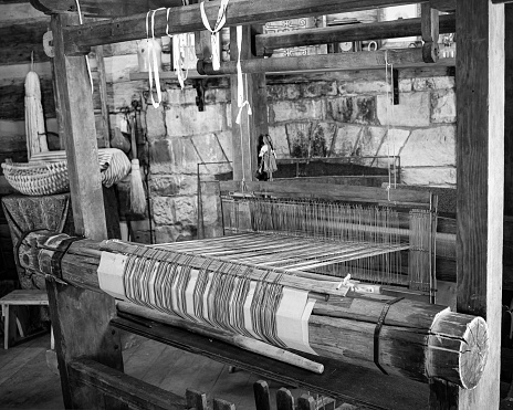 A grayscale of an old wooden loom making cloth from cotton threads in a room with stone walls