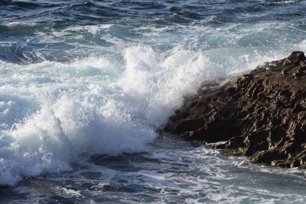 istock Closeup of sunlit water waves hitting uneven beach stone making foam 1446632355
