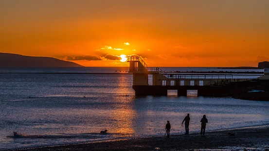 A family walks on a sandy beach near Galway Bay at the Blackrock Diving Tower during bright orange sunset