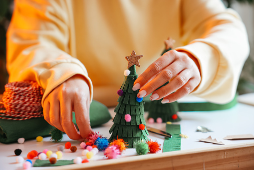 Young Woman Wrapping Christmas Gifts In Living Room. Creative diy craft hobby. Making handmade craft christmas ornaments and balls with felt spruce tree. Woman's leisure, holiday decorations. Closeup of female hands at white wood background