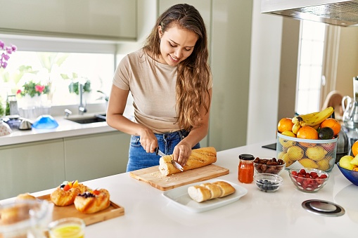 Young beautiful hispanic woman preparing breakfast cutting bread at the kitchen