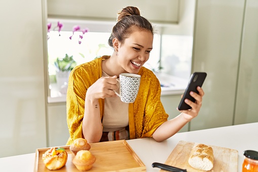 Young beautiful hispanic woman having breakfast drinking coffee using smartphone at the kitchen